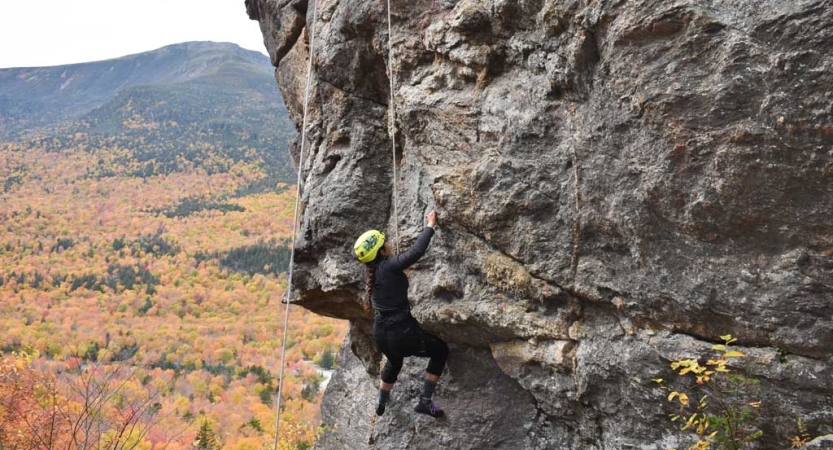 A person wearing safety gear is secured by ropes as they rock climb high above fall foliage below. 